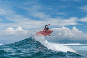 Surfer performing an aerial maneuver on a pink surfboard amidst ocean waves, representing summer sports activities related to injuries and imaging services at Life Medical Imaging.